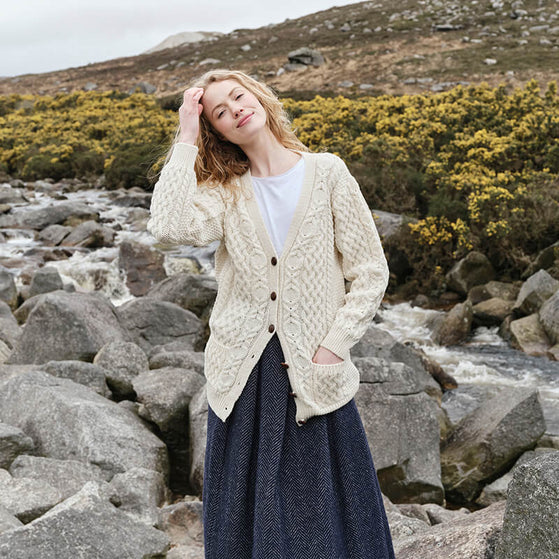 A person stands by a flowing river, wearing a long, knitted aran cardigan and a dark skirt. They have wavy hair and are playfully adjusting it. The background features rocky terrain and flowering shrubs under an overcast sky.
