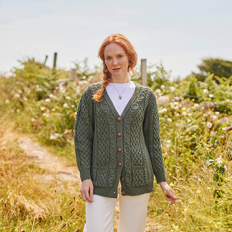 A woman stands on a path surrounded by greenery, wearing a green knitted aran cardigan with buttons over a white shirt. She has long red hair styled in a braid and looks directly at the camera. The setting features wildflowers and lush foliage in the background, suggesting an outdoor environment.