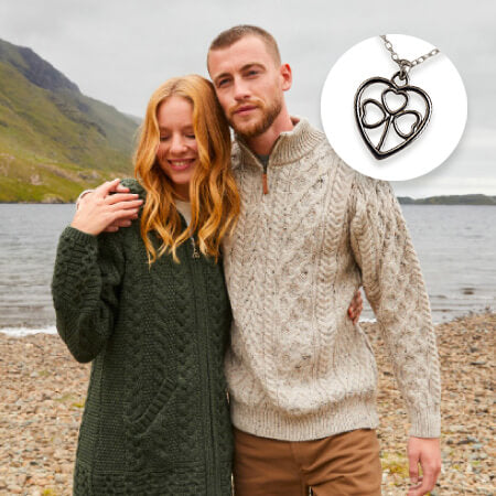 Couple standing on a pebbly beach with mountains in the background, wearing cable knit sweaters. Inset shows a necklace with a heart-shaped pendant.