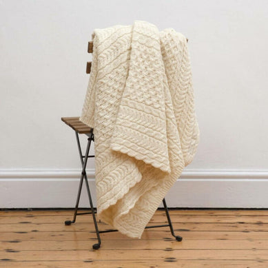 Cream cable-knit Irish blanket with diamond and Celtic-inspired patterns, draped over a rustic wooden chair on a natural wood floor—evoking traditional Aran knitting heritage and timeless Irish home décor charm.