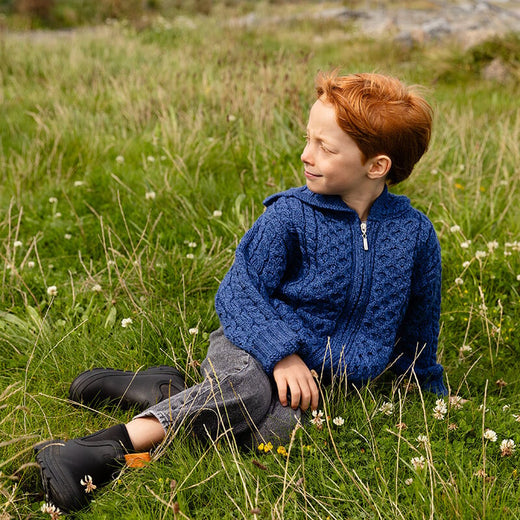 A child with red hair sits on a grassy field, wearing a blue knitted aran kids sweater and gray pants. The scene is natural, with green grass and scattered wildflowers in the background. The child looks off to the side, appearing relaxed and contemplative.