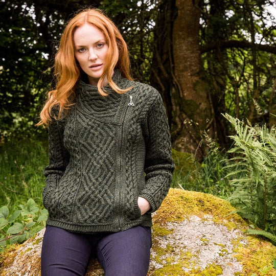 A woman with long red hair sits on a moss-covered rock in a forested area. She is wearing a dark green, textured sweater with a high collar and two front pockets. The background features trees and greenery, creating a natural setting.