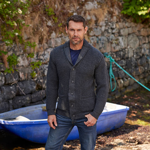 A man stands in front of a stone wall near a blue boat. He wears a dark gray, buttoned Aran cardigan over a navy shirt and has an attentive expression. The setting appears outdoors with some greenery in the background.