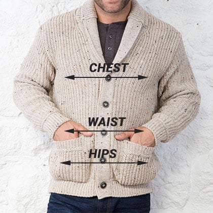 A man wearing a beige, knitted aran cardigan stands against a textured white wall. He has his hands in the pockets of the sweater. Text labels indicate measurements for chest, waist, and hips on the garment.