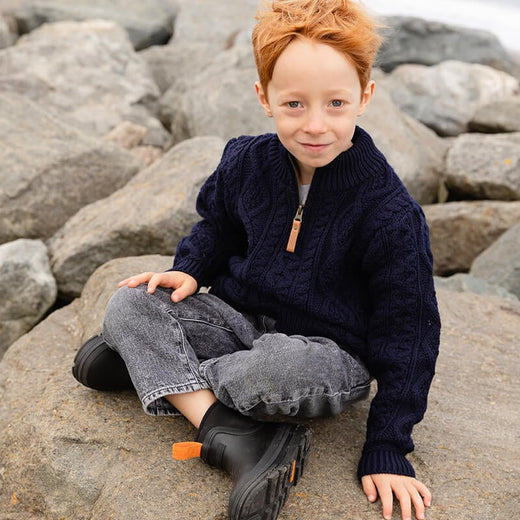 A young boy with curly red hair sits on large rocks by the water. He wears a dark navy kids Aran sweater and gray jeans, paired with black shoes. The background is coastal, suggesting an outdoor setting.