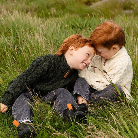 Two young children with red hair are sitting on a grassy field, smiling and playfully leaning toward each other. One child wears a dark green kids Aran sweater and the other a cream-colored merino wool kids sweater. Both are dressed in denim jeans. The scene captures a moment of joy and friendship in a natural setting.