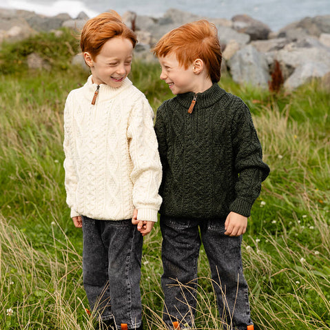 Two young boys stand together in a grassy field. One boy wears a cream-colored kids Aran sweater, while the other is dressed in a dark green sweater. They are smiling at each other, with rocky terrain and a distant ocean in the background.