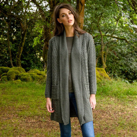 A woman stands outdoors in a green setting, wearing a long, green knitted aran cardigan over a gray top and blue jeans. She gazes at the camera with a neutral expression.