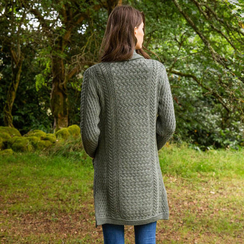 A woman stands with her back to the camera, wearing a long, green knitted aran cardigan. The background features lush greenery and trees, creating a natural setting. The focus on her attire highlights the textured pattern of the cardigan.