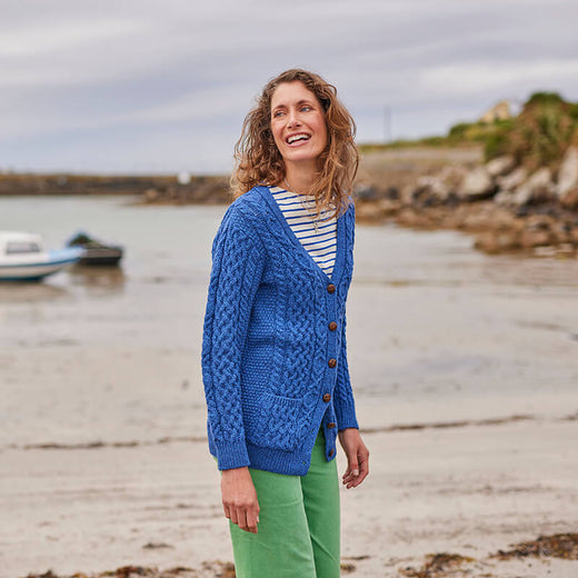 A woman stands on a beach, smiling. She is wearing a blue knitted aran cardigan over a striped shirt and green pants. In the background, there is a small boat and rocky shoreline under a cloudy sky.