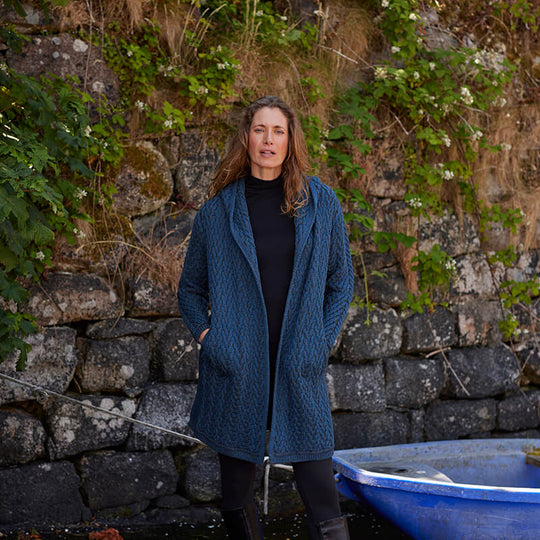 Womens Signature Hooded Aran Waterfall Cardigan in blue with textured knit, worn outdoors near a stone wall and blue boat.