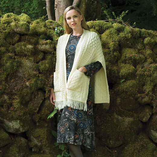 A woman stands against a moss-covered stone wall, wearing a floral-patterned dress and a light-colored knitted aran shawl with pockets. She has straight hair and appears relaxed in the natural setting.