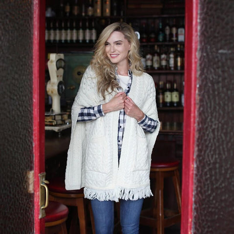 Cream merino wool pocket shawl with cable knit patterns and fringe, worn over a plaid shirt while standing in a pub doorway.
