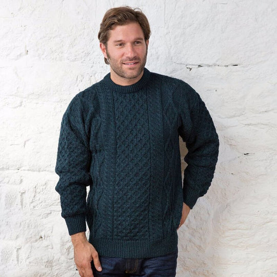 A man stands against a white stone wall, wearing a dark blue knitted aran sweater with a textured pattern. He has short brown hair and is smiling slightly while posing with one hand in his pocket.