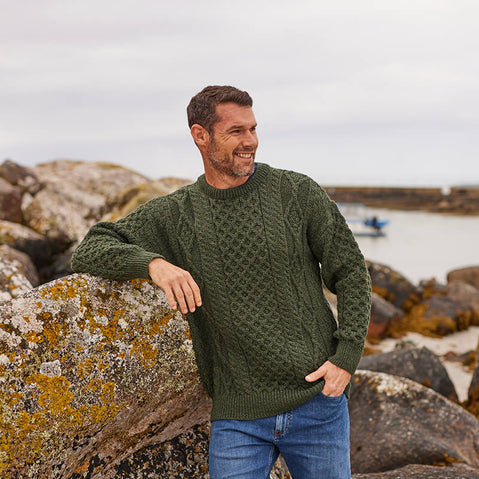 A man stands on a rocky beach, wearing an olive green knitted aran sweater. He is smiling and leaning against a large rock, with a calm sea and boat visible in the background under a cloudy sky.