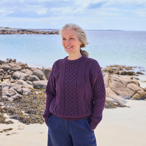 A woman stands on a beach, smiling with her hands in her pockets. She wears a purple, textured aran sweater and loose-fitting blue pants. The background features calm water and rocky shorelines under a partly cloudy sky.