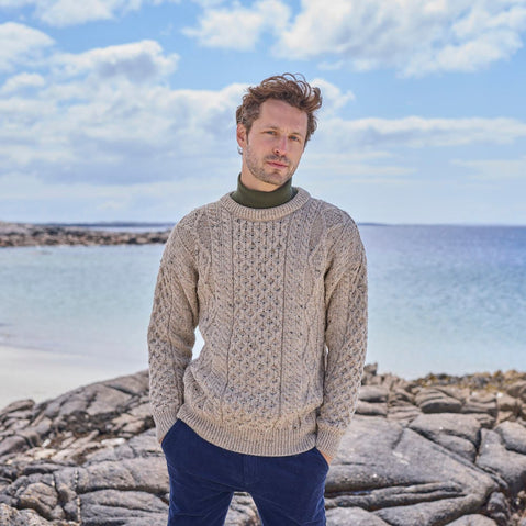 A man stands on rocky beach terrain, wearing a light gray cable-knit aran sweater over a dark turtleneck. He gazes thoughtfully into the distance, with calm blue water and a cloudy sky in the background.