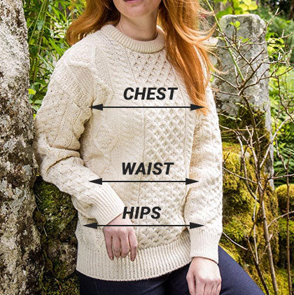 A person wearing a cream-colored cable-knit aran sweater stands outdoors, surrounded by greenery and rocks. Text labels indicate measurement points for
