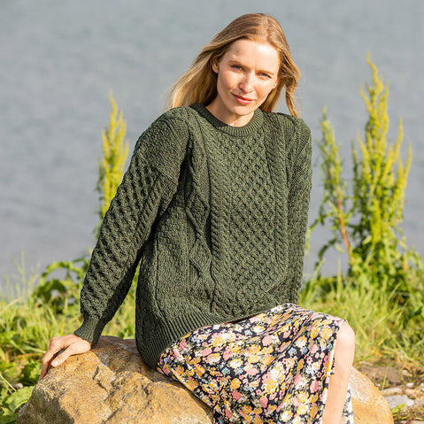Woman wearing an army green Fisherman Lightweight Crew Neck Heritage Sweater with a textured knit pattern, paired with a floral skirt, sitting on a rock with greenery and water in the background.