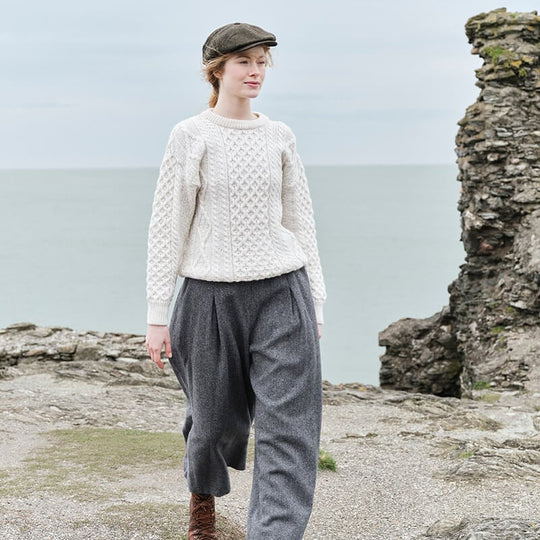A woman walks along a rocky shoreline, wearing a white cable-knit aran sweater, gray wide-leg trousers, and a green cap. The ocean is visible in the background under an overcast sky.