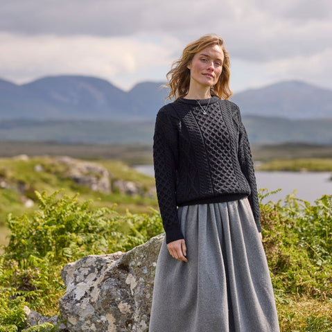 A woman stands outdoors in a scenic landscape, wearing a black knitted aran sweater and a flowing gray skirt. She has wavy hair and appears relaxed, with mountains and greenery in the background. The sky is partly cloudy, suggesting a mild day.