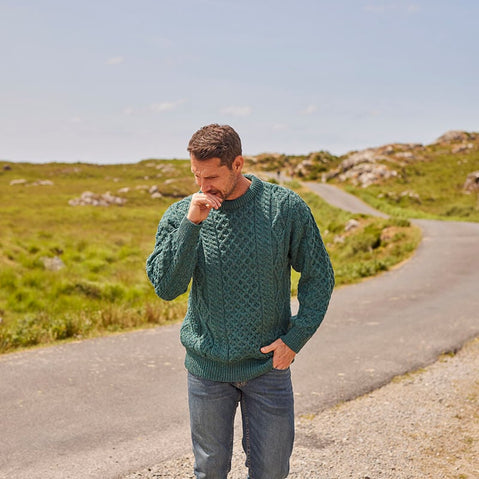 A man in a green cable-knit aran sweater stands on a gravel path, gently touching his chin with one hand. He is surrounded by a scenic landscape of rolling hills and sparse vegetation under a clear sky.