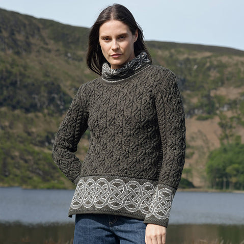 Woman wearing a dark green knitted Aran sweater with Celtic patterns standing in front of a scenic landscape.