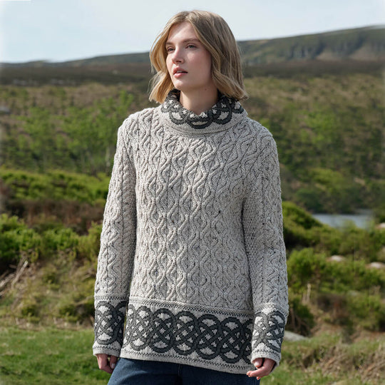 Woman wearing a grey patterned Aran sweater standing in a scenic outdoor setting with hills and water.