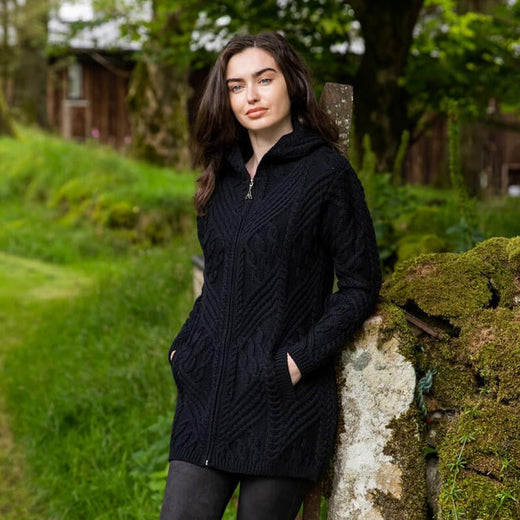 A young woman in a black zip-up hooded Aran sweater stands beside a moss-covered stone, with lush green grass and trees in the background. She has a neutral expression and her hands are in the pockets of her sweater. Soft natural light highlights the scene.