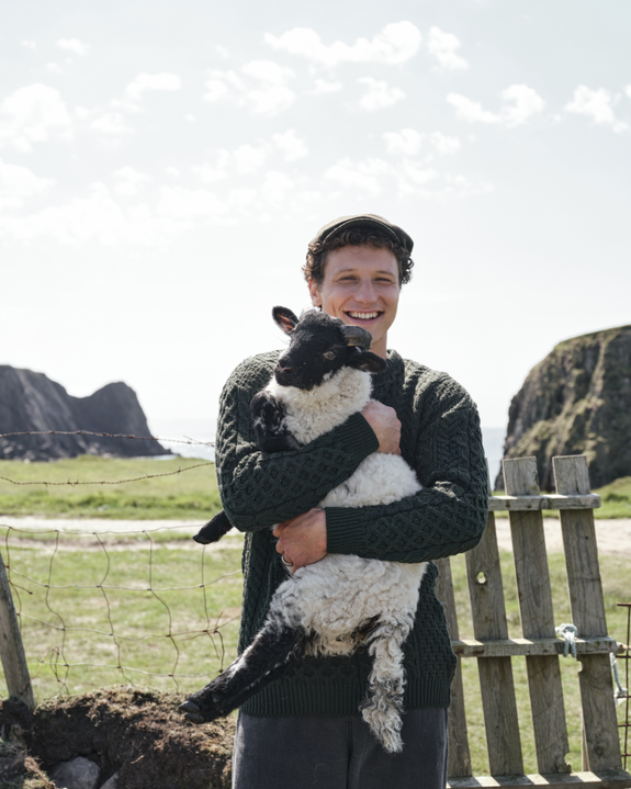 A man in a green sweater smiling while holding a black and white lamb against a scenic backdrop of cliffs and fields.