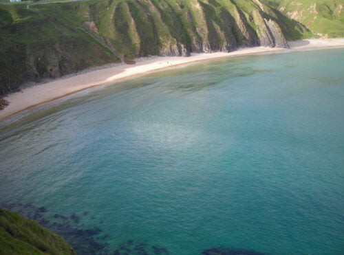Aerial view of a tranquil Donegal beach with soft, sandy shorelines surrounded by lush green cliffs. The clear blue water gently laps at the coast, reflecting the sunlight.