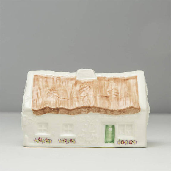 A Belleek china model of a cottage with a textured brown roof and white walls. The front features a small green door and decorative flowers below the windows. The background is plain and neutral.