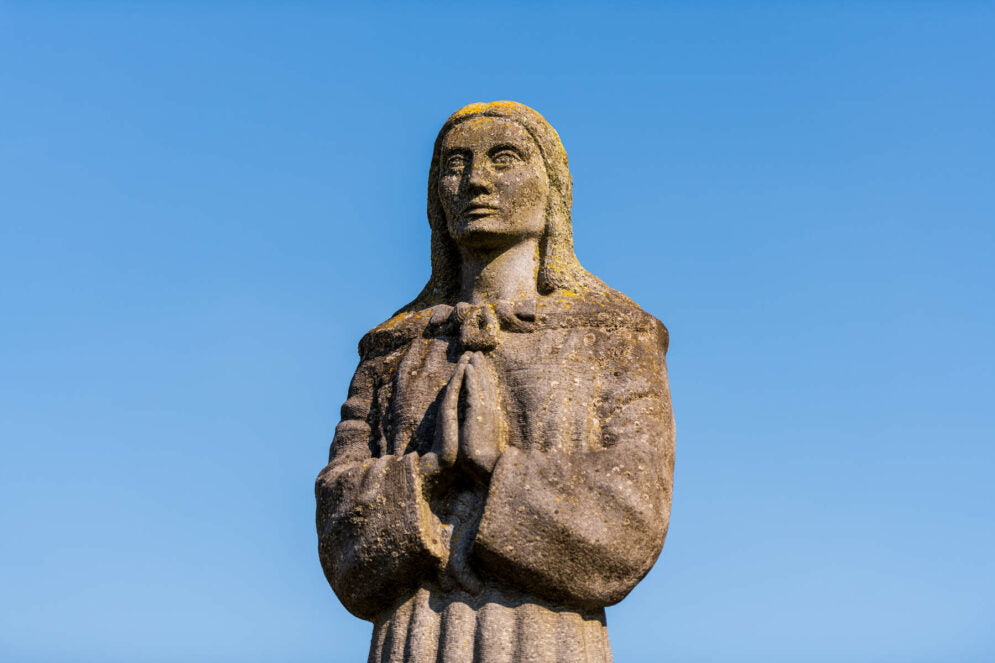 A stone statue of Saint Brigid, with long hair and clasped hands, set against a clear blue sky. The figure wears a simple garment and has a serene expression.