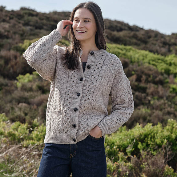 Person wearing a beige Women's Traditional Cable Knit Aran Cardigan, featuring intricate cable knit patterns and dark buttons