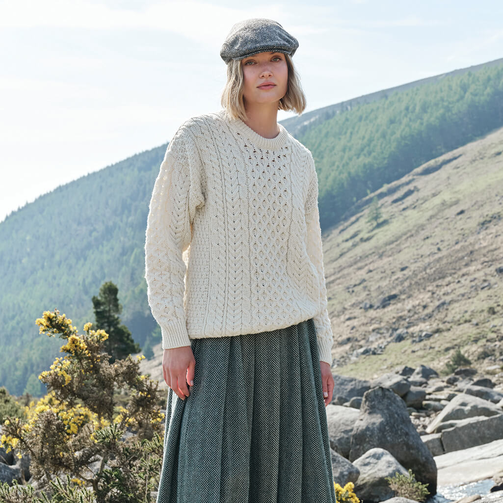 Woman wearing a cream Aran sweater and green skirt standing in a mountainous landscape.