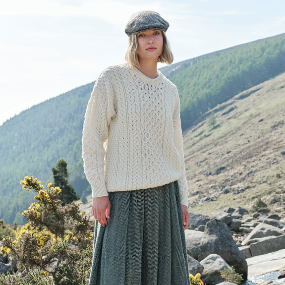 Woman wearing a cream Aran sweater and green skirt standing in a mountainous landscape.