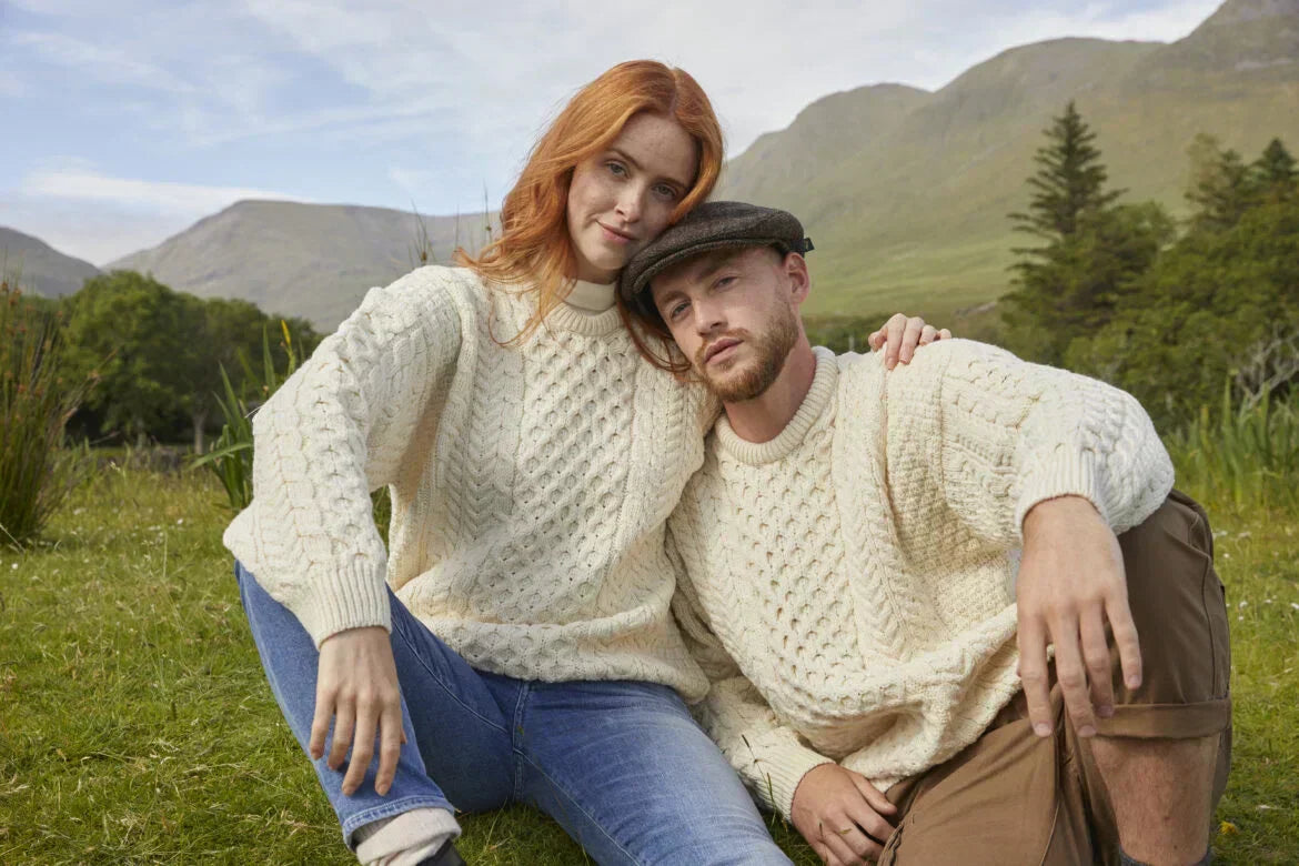 A man and a woman sit on grassy terrain, both wearing cream-colored cable-knit Aran sweaters. In the background, green hills and trees are visible under a blue sky.