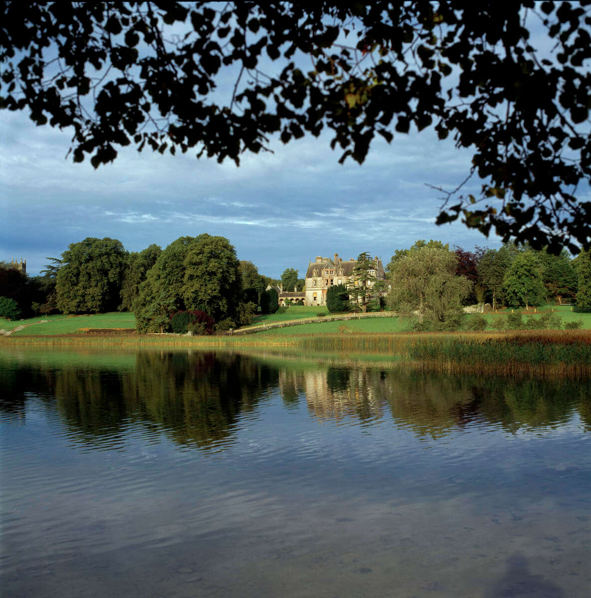 A picturesque view of a stately manor house set against a backdrop of lush trees, with a calm lake in the foreground reflecting the building and foliage. The sky is partly cloudy, adding depth to the scene.