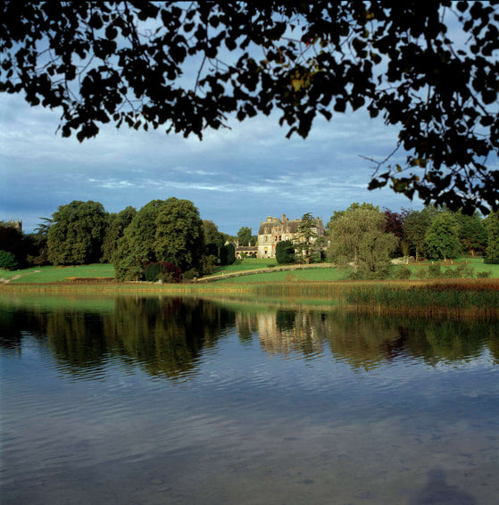 A picturesque view of a stately manor house set against a backdrop of lush trees, with a calm lake in the foreground reflecting the building and foliage. The sky is partly cloudy, adding depth to the scene.