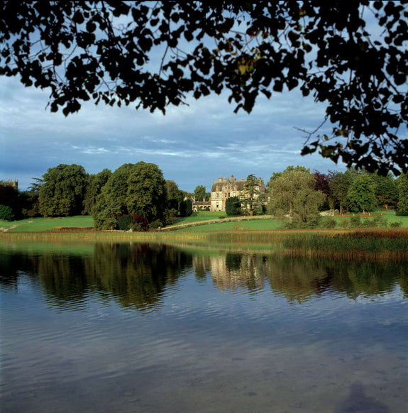 A picturesque view of a stately manor house set against a backdrop of lush trees, with a calm lake in the foreground reflecting the building and foliage. The sky is partly cloudy, adding depth to the scene.