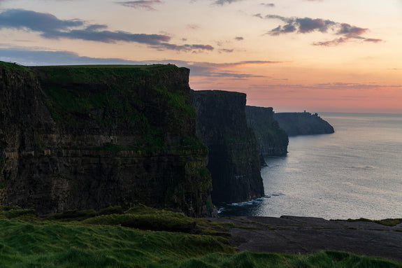Cliffs of Moher, County Clare. Dramatic cliffs rise sharply from the ocean at sunset, with a soft glow in the sky illuminating the rugged coastline.