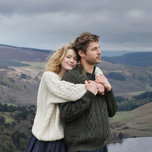 A couple in matching knit sweaters embraces against a backdrop of rolling hills and a gray sky.