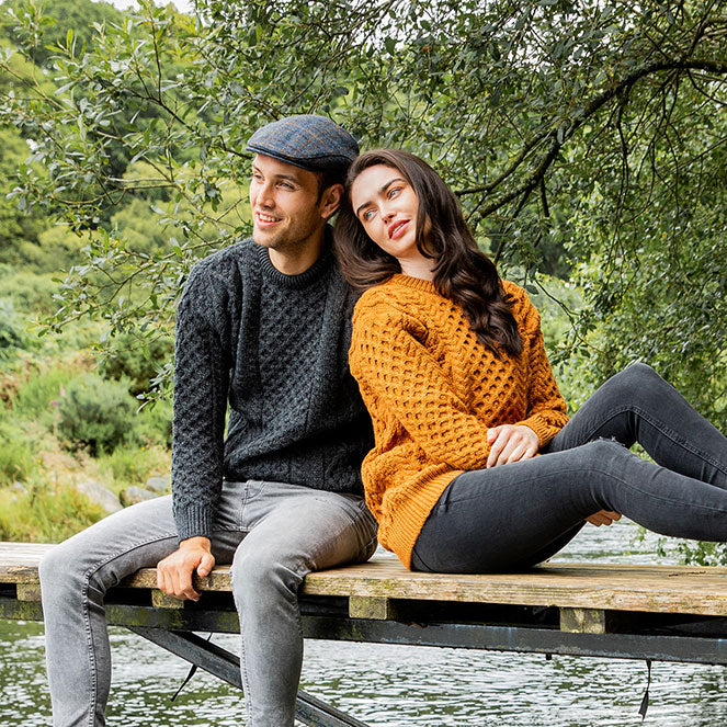 A man in a dark Aran sweater and flat cap sits on the edge of a wooden dock beside a woman in an orange Aran sweater. They are both smiling, with the backdrop featuring lush greenery and a calm body of water.