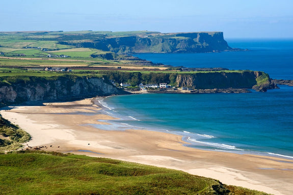 A scenic view of an Irish coastline featuring a sandy beach and clear blue waters. Cliffs rise in the background, surrounded by green fields under a bright sky. 
