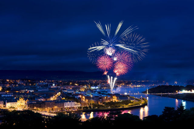 Fireworks illuminate the night sky above a cityscape, with vibrant colors reflecting off a river. 