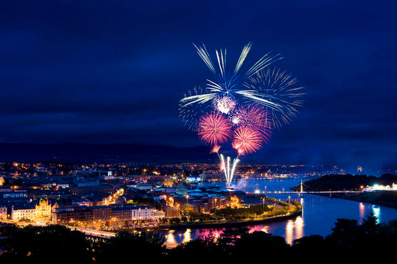 Fireworks illuminate the night sky above a cityscape, with vibrant colors reflecting off a river. 