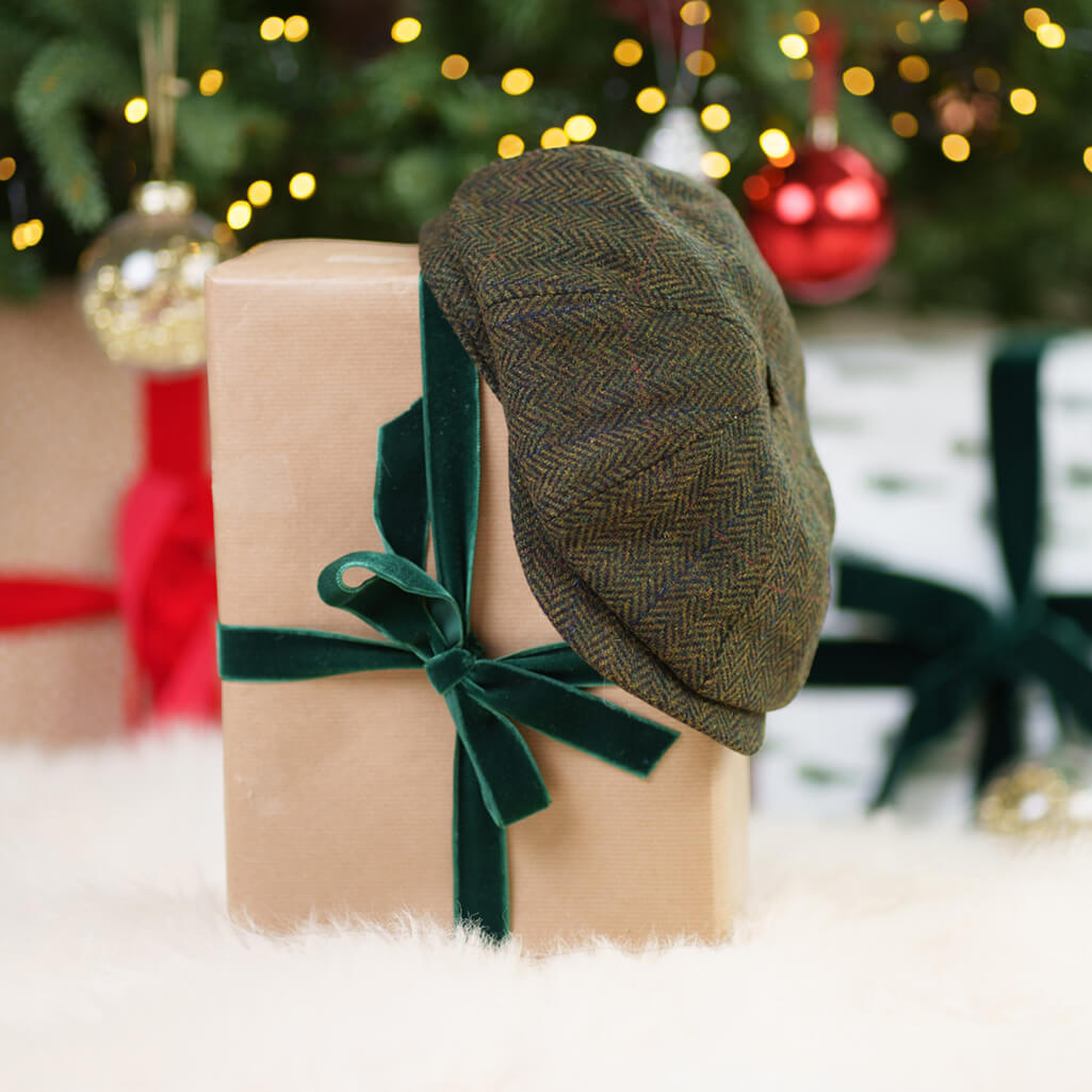 Olive green Irish tweed flat cap with herringbone pattern resting on kraft paper gift box tied with dark green velvet ribbon, surrounded by Christmas decorations and bokeh lights.