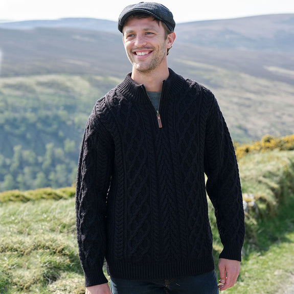A man wearing a black knitted sweater and a flat cap stands smiling in a hilly landscape with green grass and distant mountains.