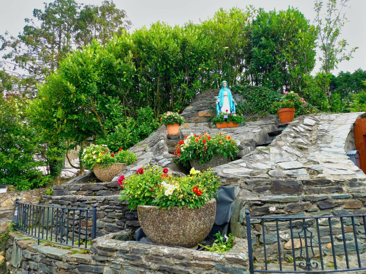 A rocky garden featuring a statue of the Virgin Mary, surrounded by vibrant flower pots. Lush greenery frames the scene.