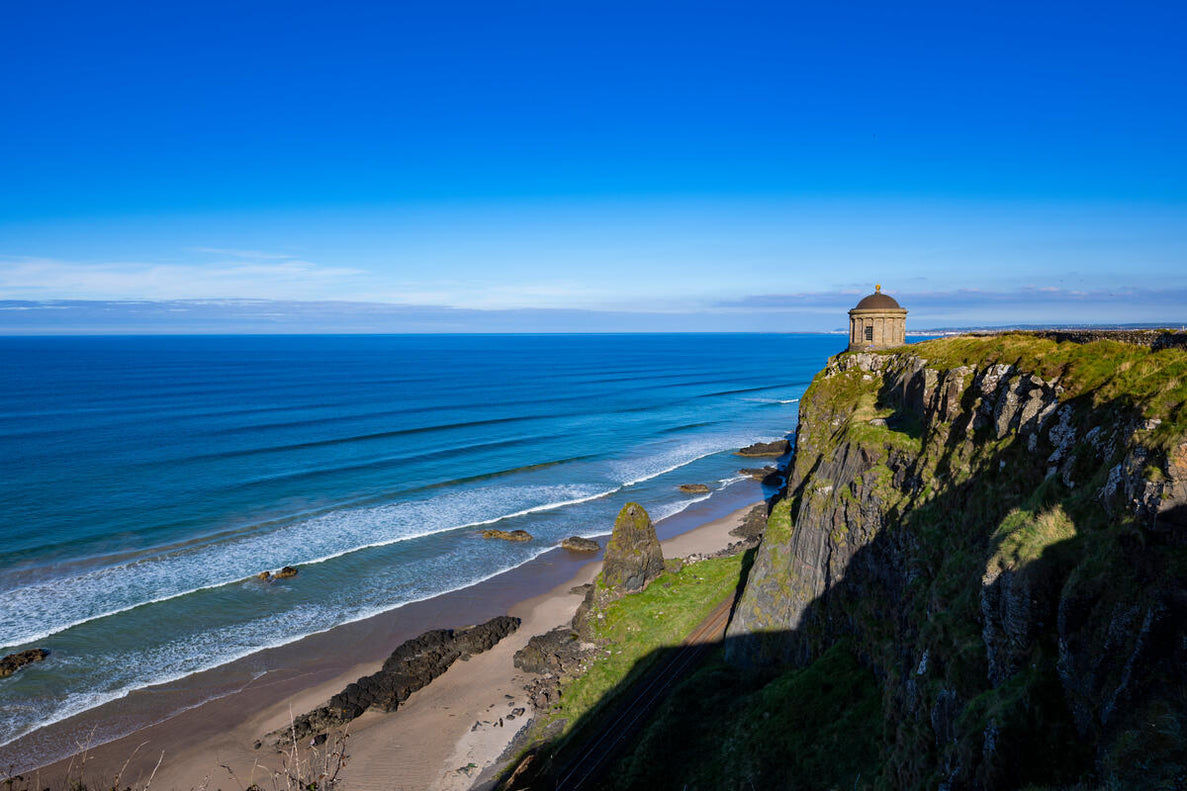 A coastal cliffside with a small, cylindrical stone building perched on the edge, overlooking a vast ocean. Waves gently roll onto a sandy beach below, under a clear blue sky.